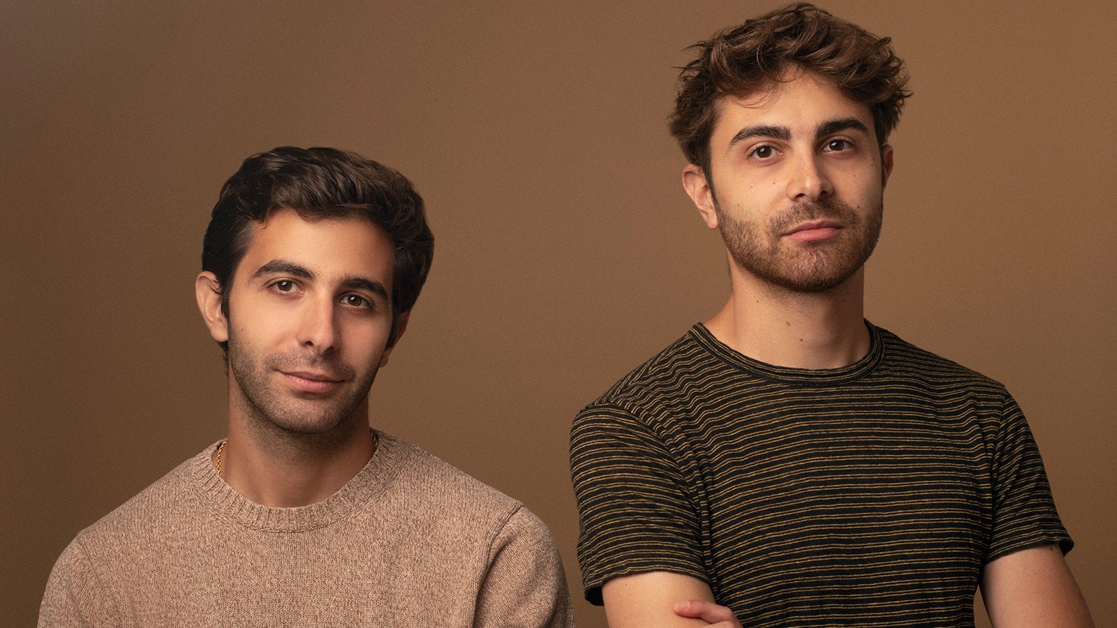 Two men with short brown hair wearing t-shirts in front of a light brown background