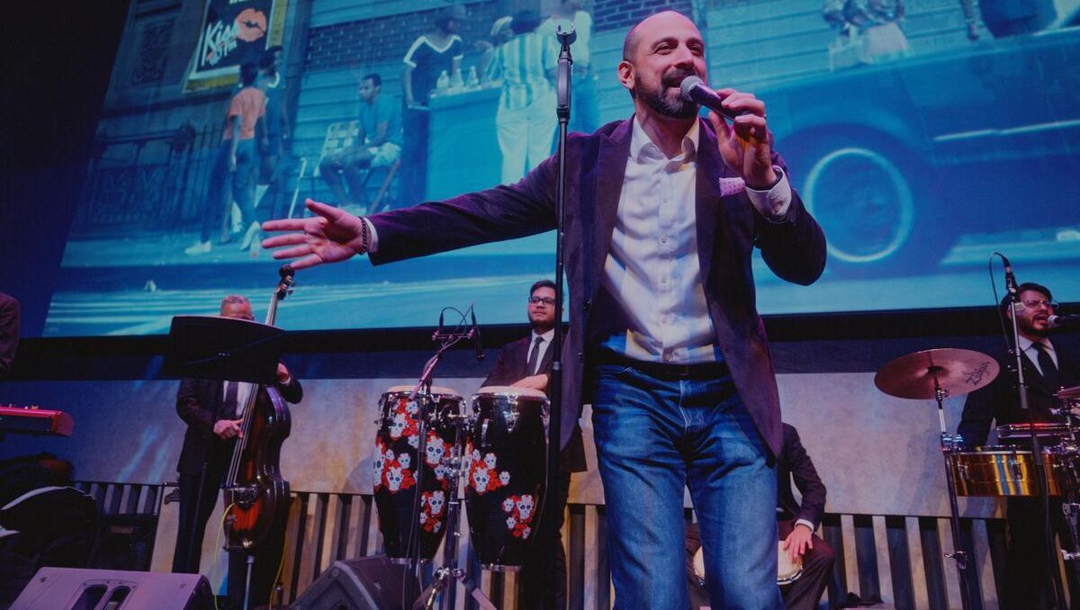 Man in a blazer, jeans, and white button down singing on stage