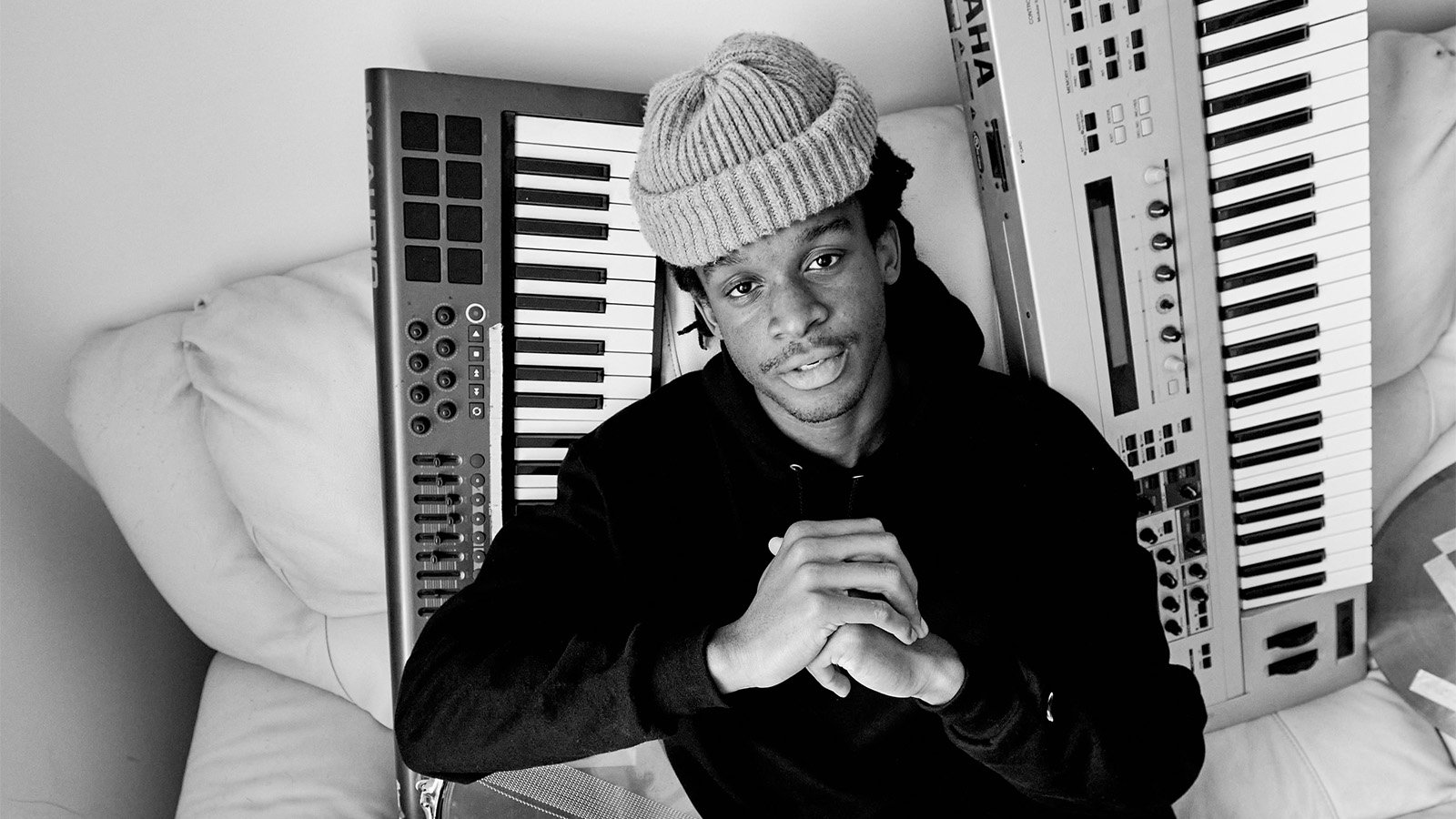 Black and white photo of a man in a beanie sitting in front of two keyboards