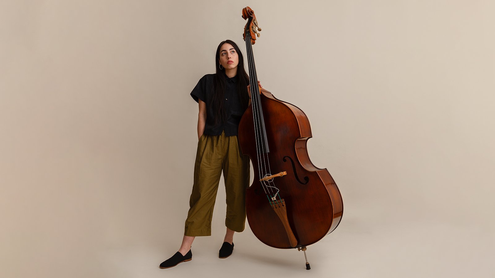 A woman with long brown hair wearing a black shirt and green pants holding a cello