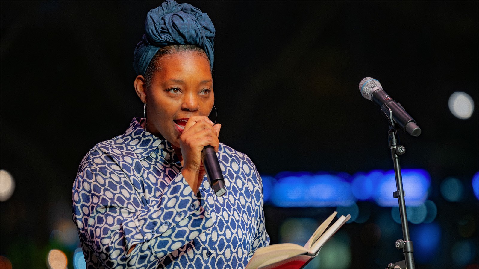A woman in a headwrap and patterned shirt reads poetry into a mic