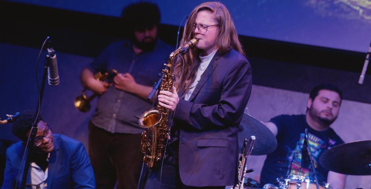 Woman in a blazer, black pants, and white turtle neck playing saxophone on stage