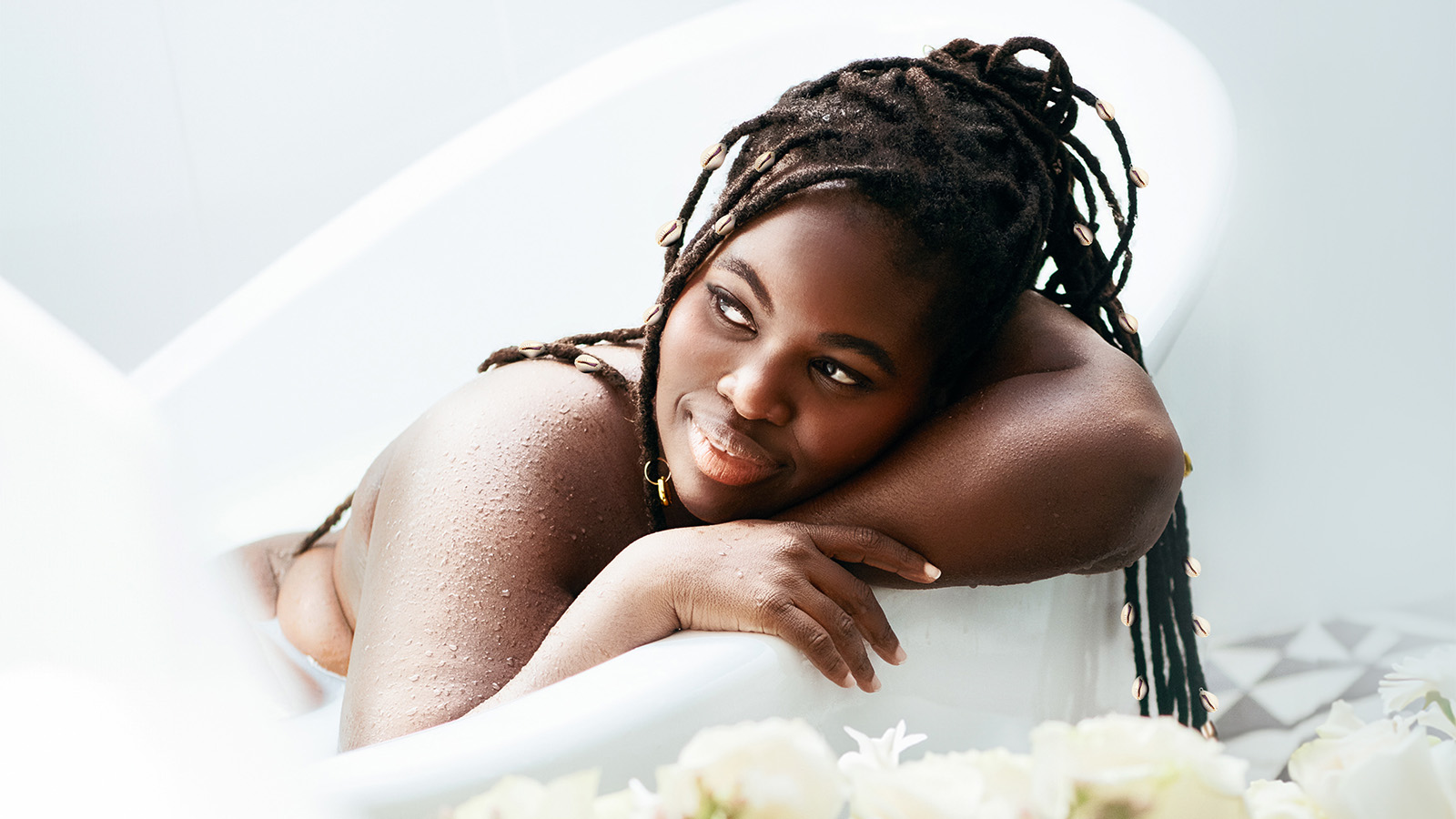 A black woman with long brown dreads laying in a white bathtub 