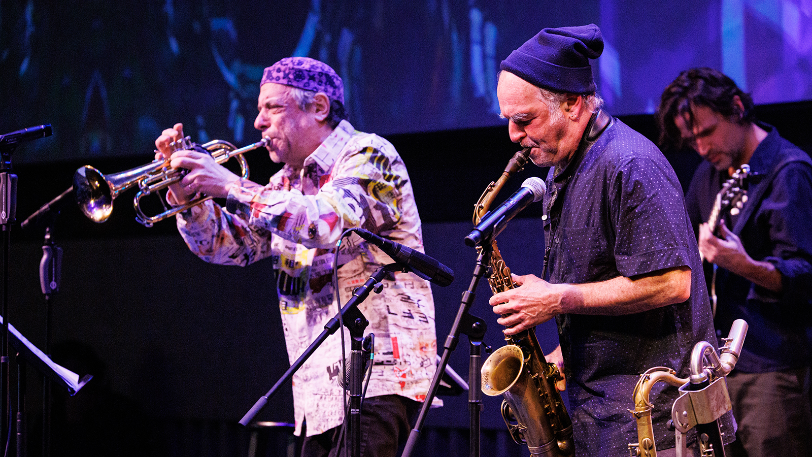 Three musicians playing brass instruments and guitar on stage.