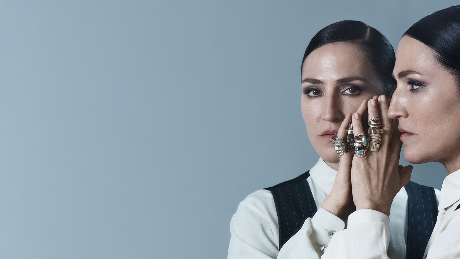 A woman with slicked back black hair holding her hand up to a mirrored version of herself