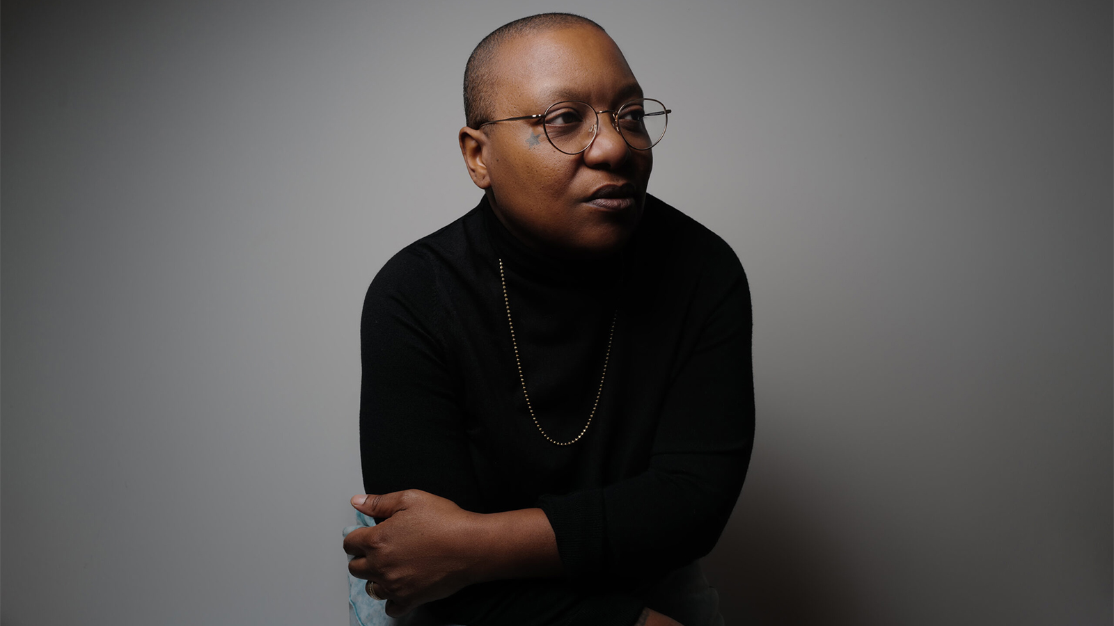 A black person wearing black clothing standing in front of a grey wall