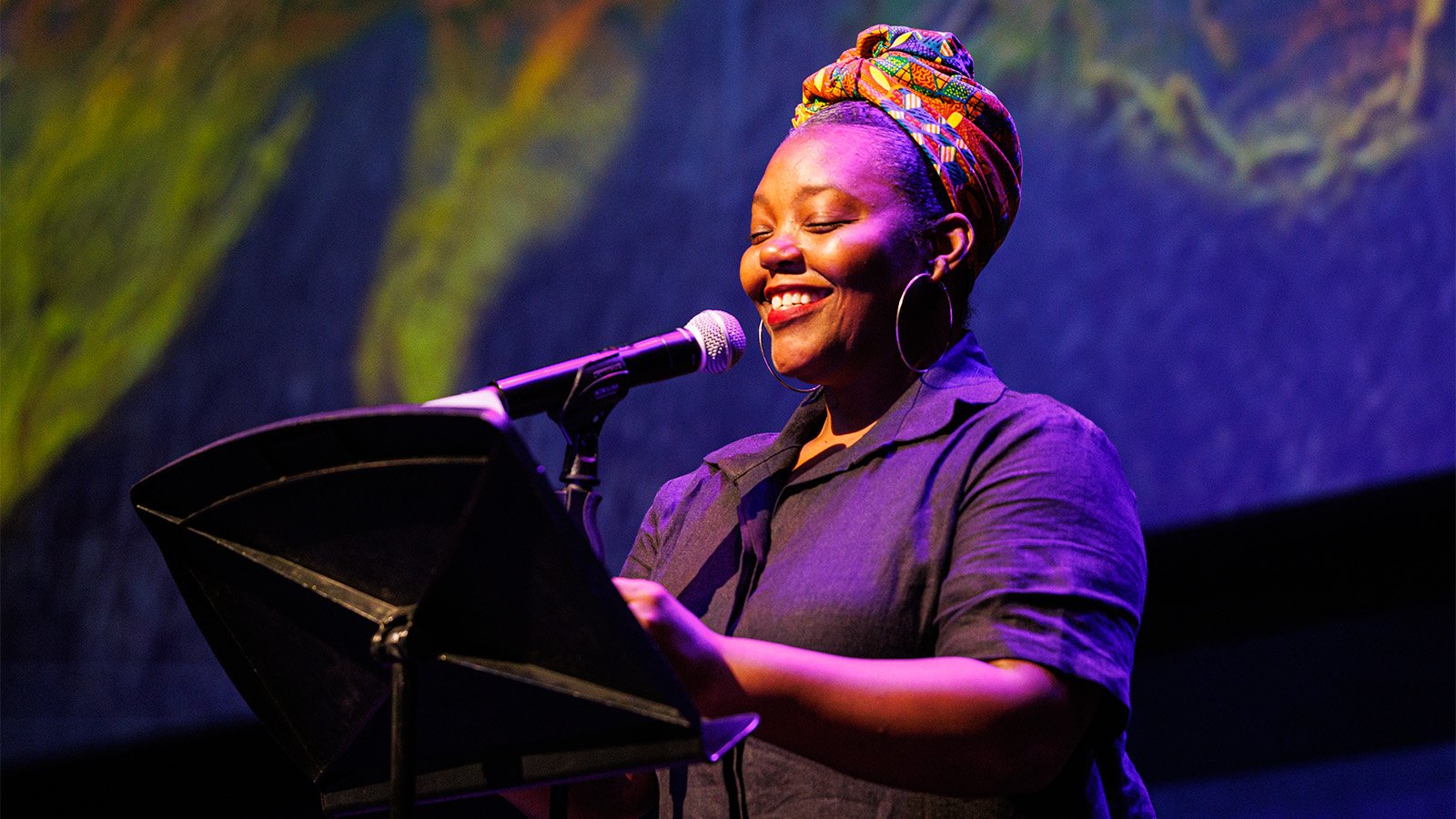 A black woman wearing a colorful head wrap standing at a microphone