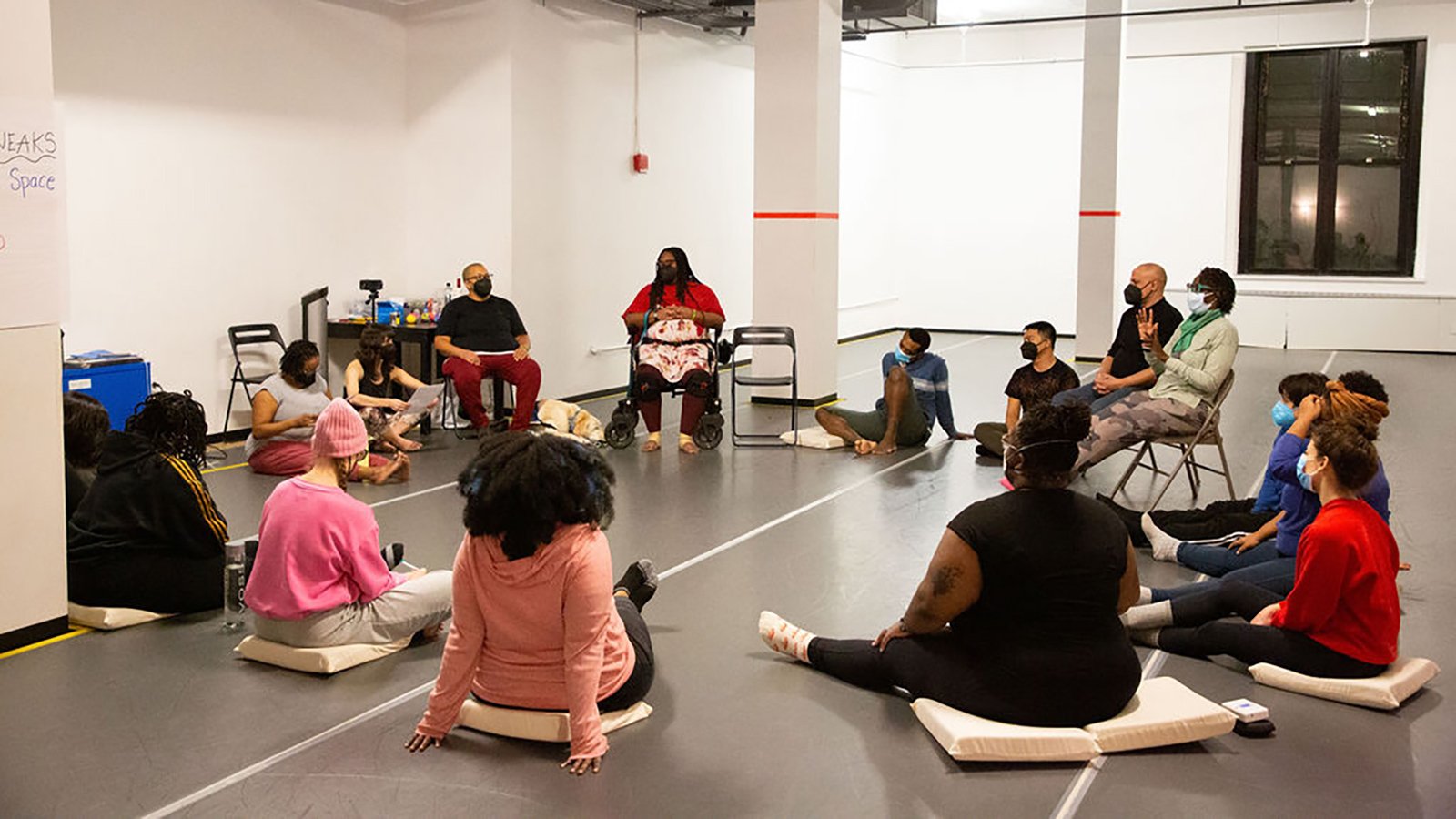 A group of disabled and non-disabled people sit in a circle with Kayla Hamilton and Elisabeth Motley. They are gathered in a dance studio with a gray floor and white walls.