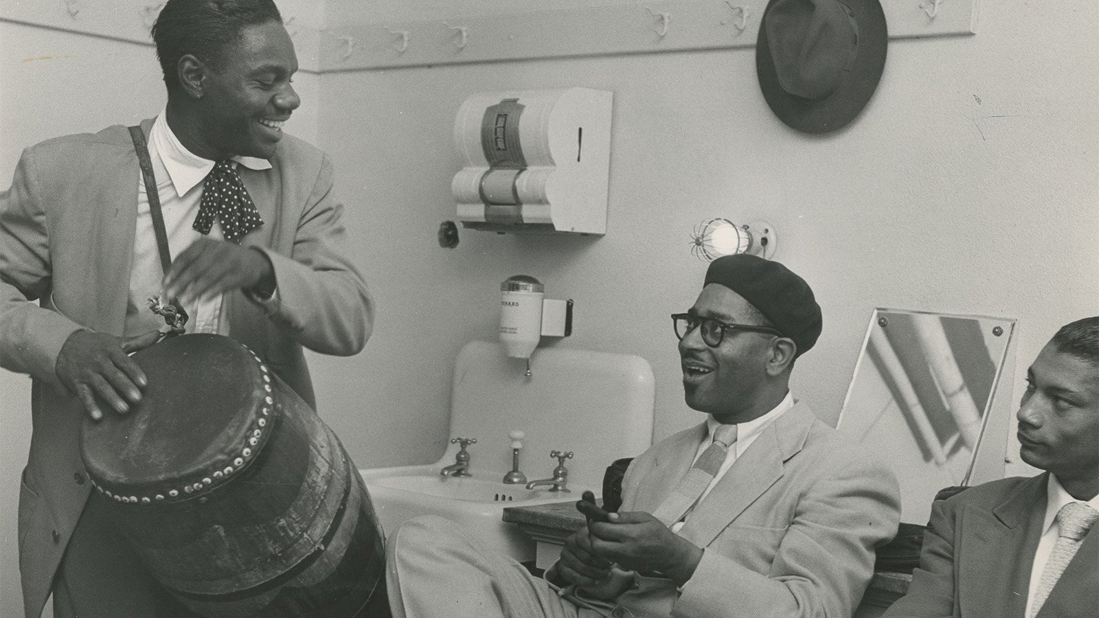A black and white photograph of a Black man playing a drum while two Black men watch. They are all in suits and ties.