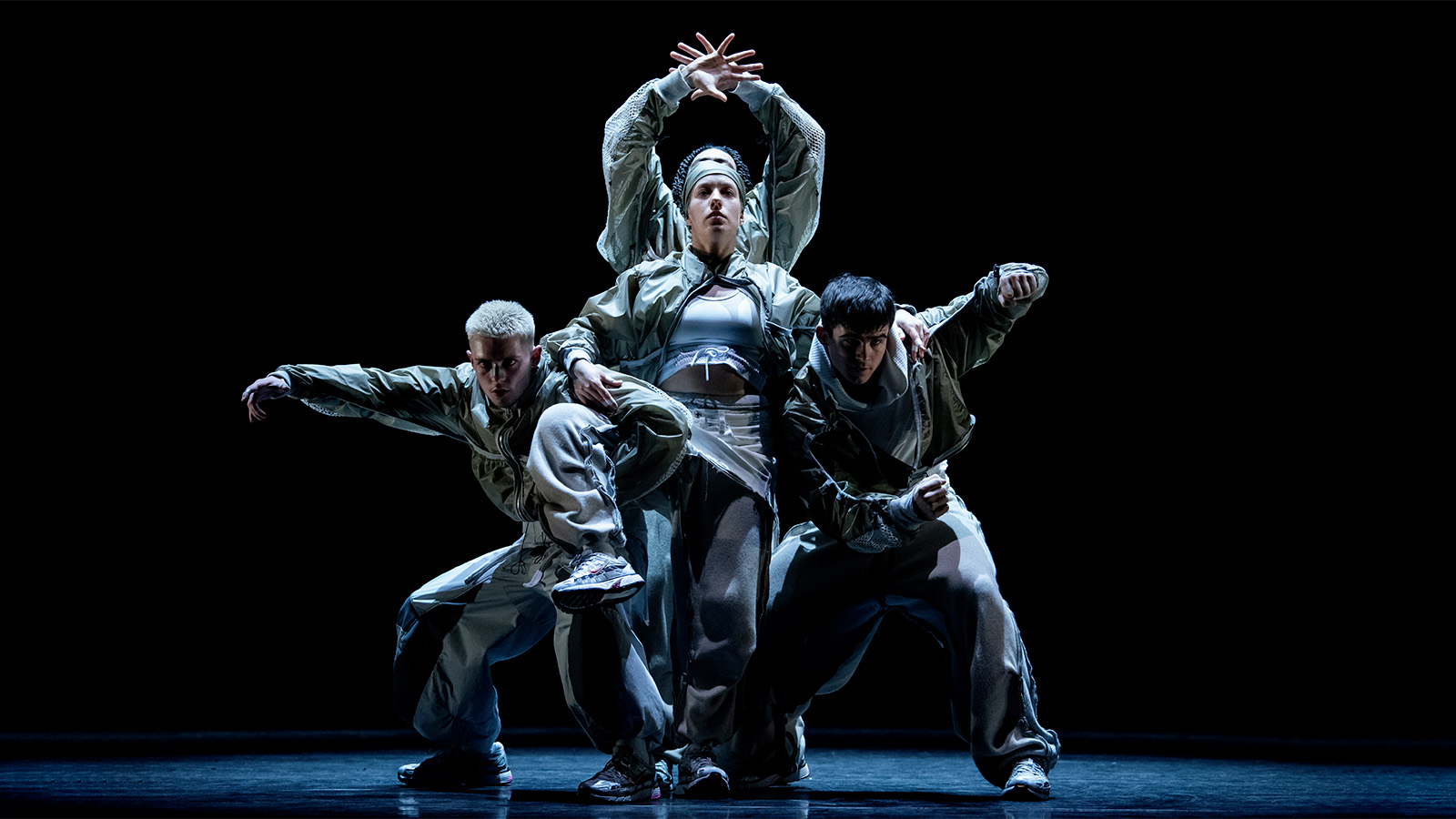Four dancers on a dark stage holding each other while looking out to the audience