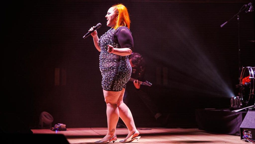 A woman with blond hair and a sparkly dress sings under a spotlight