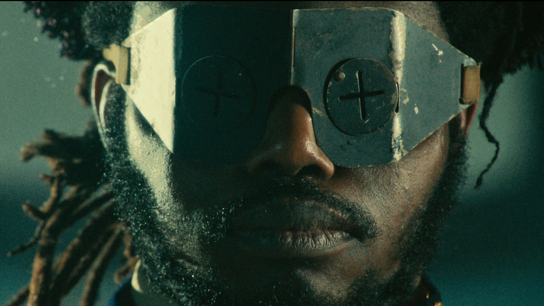 A close up image of a black man with dreads wearing a metal face covering
