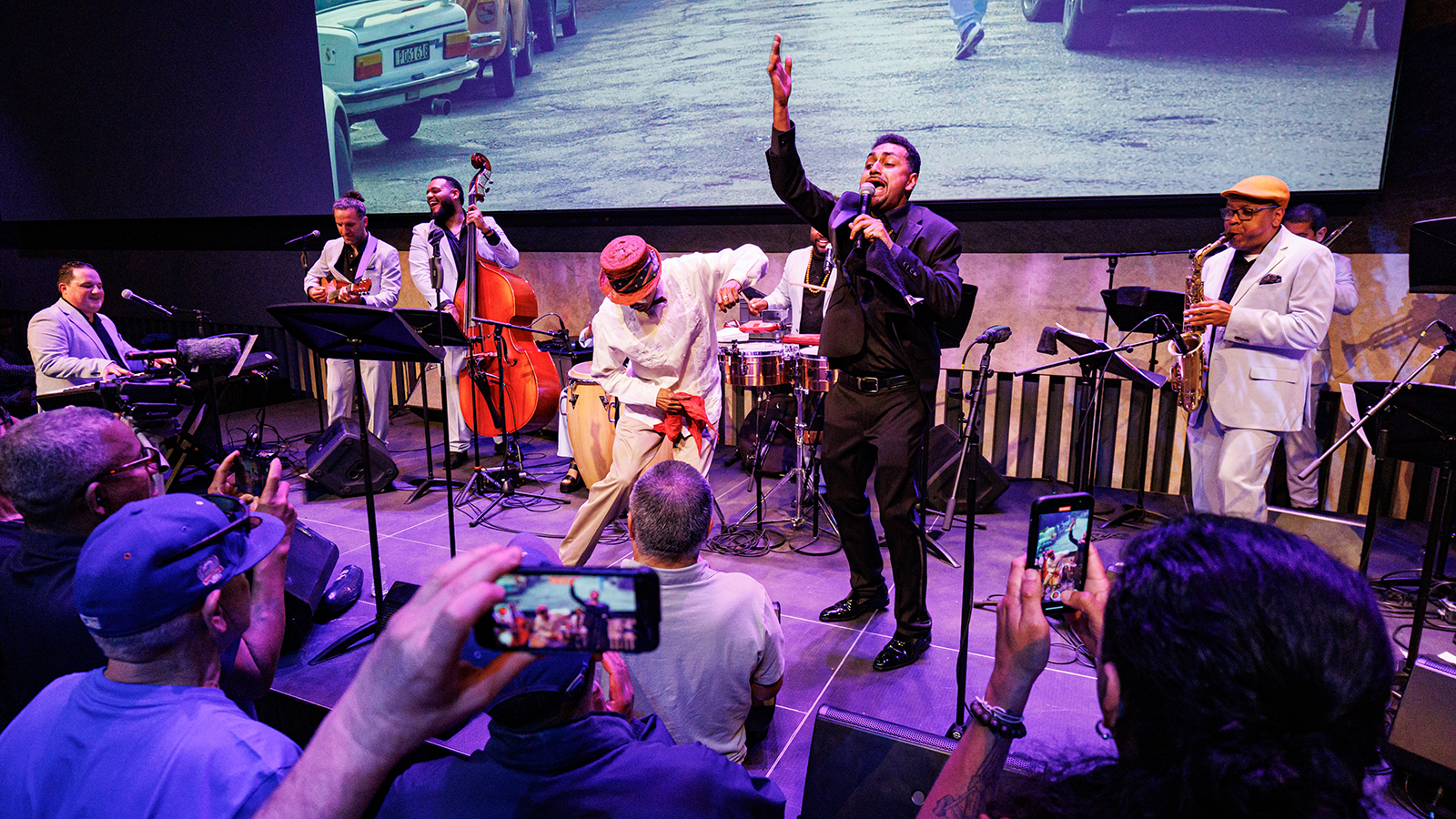 A band performs on a stage as audience members watch and film on their phones.