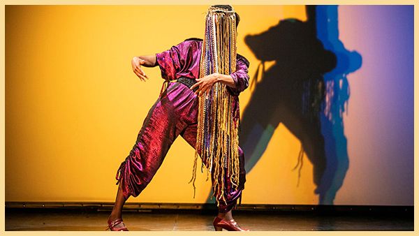 A dancer in a pink suit and a fringe face covering poses.