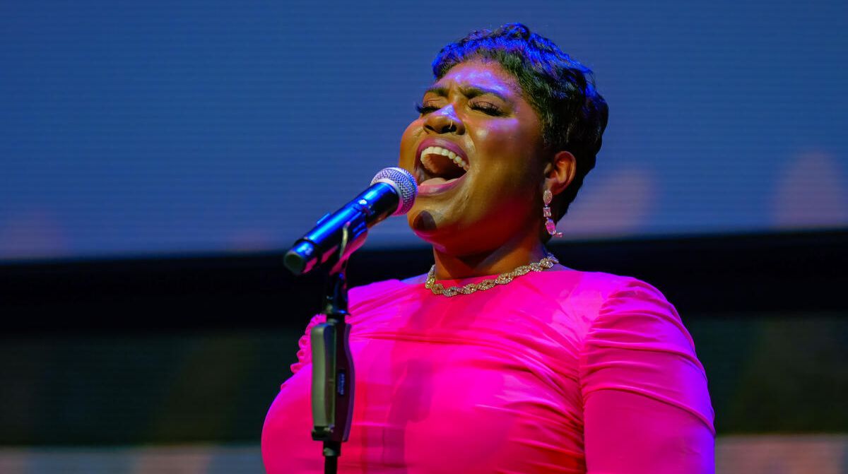 A woman with a pixie cut sings into the mic, wearing a bright pink dress and dazzling jewellery.