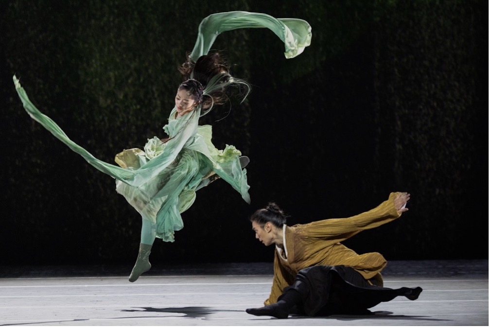 A dancer in a flowy green costume jumps in the air as another dancer bends low to the ground next to her