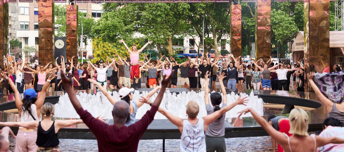 A crowd of people stand around Lincoln Center's fountain and raise their arms