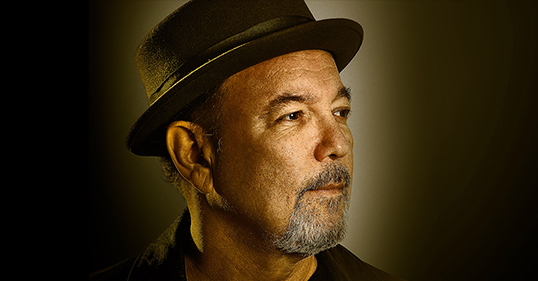 Headshot of Ruben Blades wearing a black jacket and shirt and hat and looking off to the right