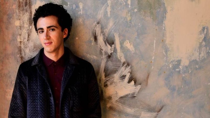 A man poses in a black blazer against a marble wall
