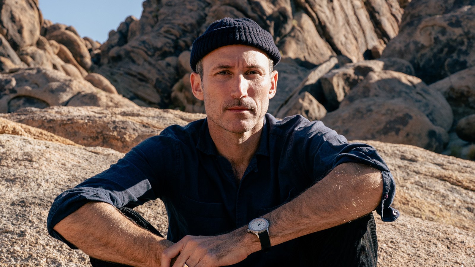 Matt Costa poses sitting down with his arms over his knees, against a rocky background, wearing a beanie and navy shirt