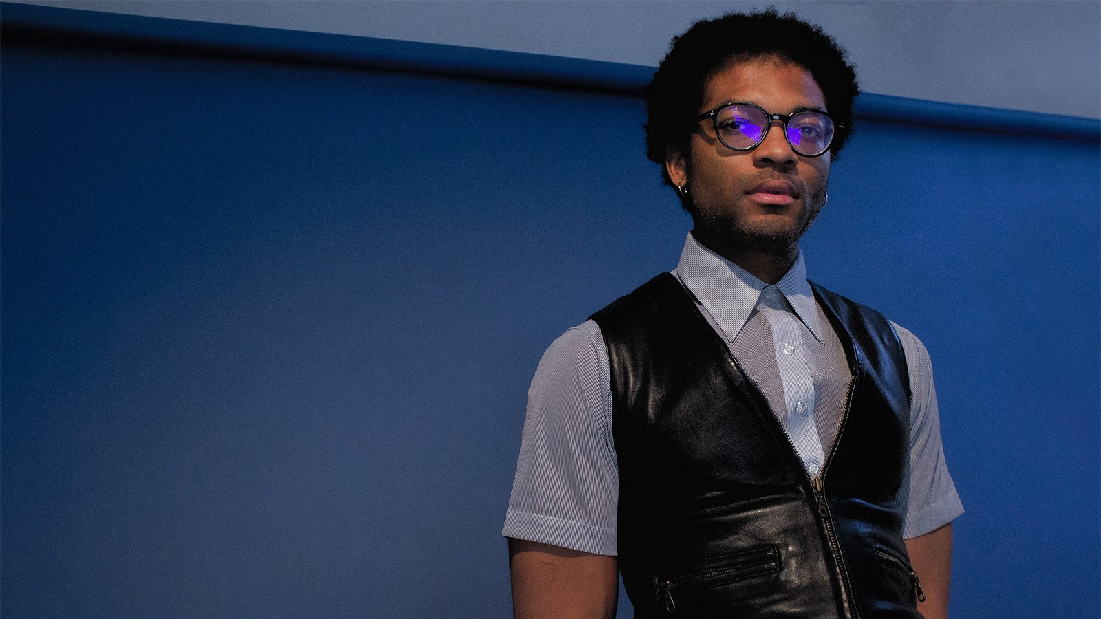 An image of Fusilier in a leather vest and a polo standing against a blue wall