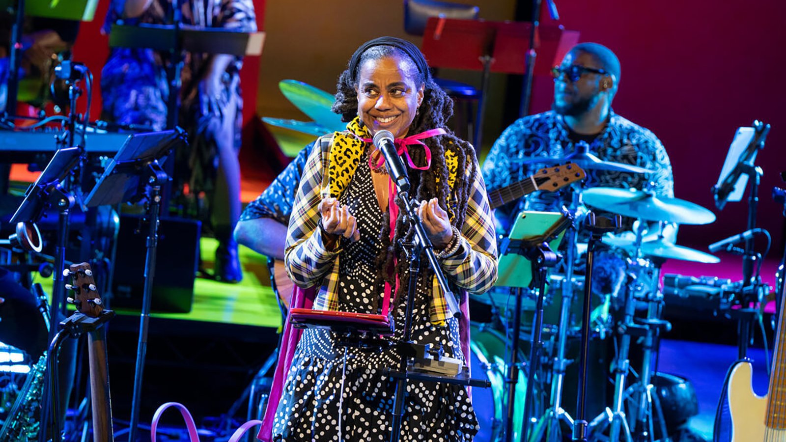 A woman in festive attire stands in front of a mic and a band
