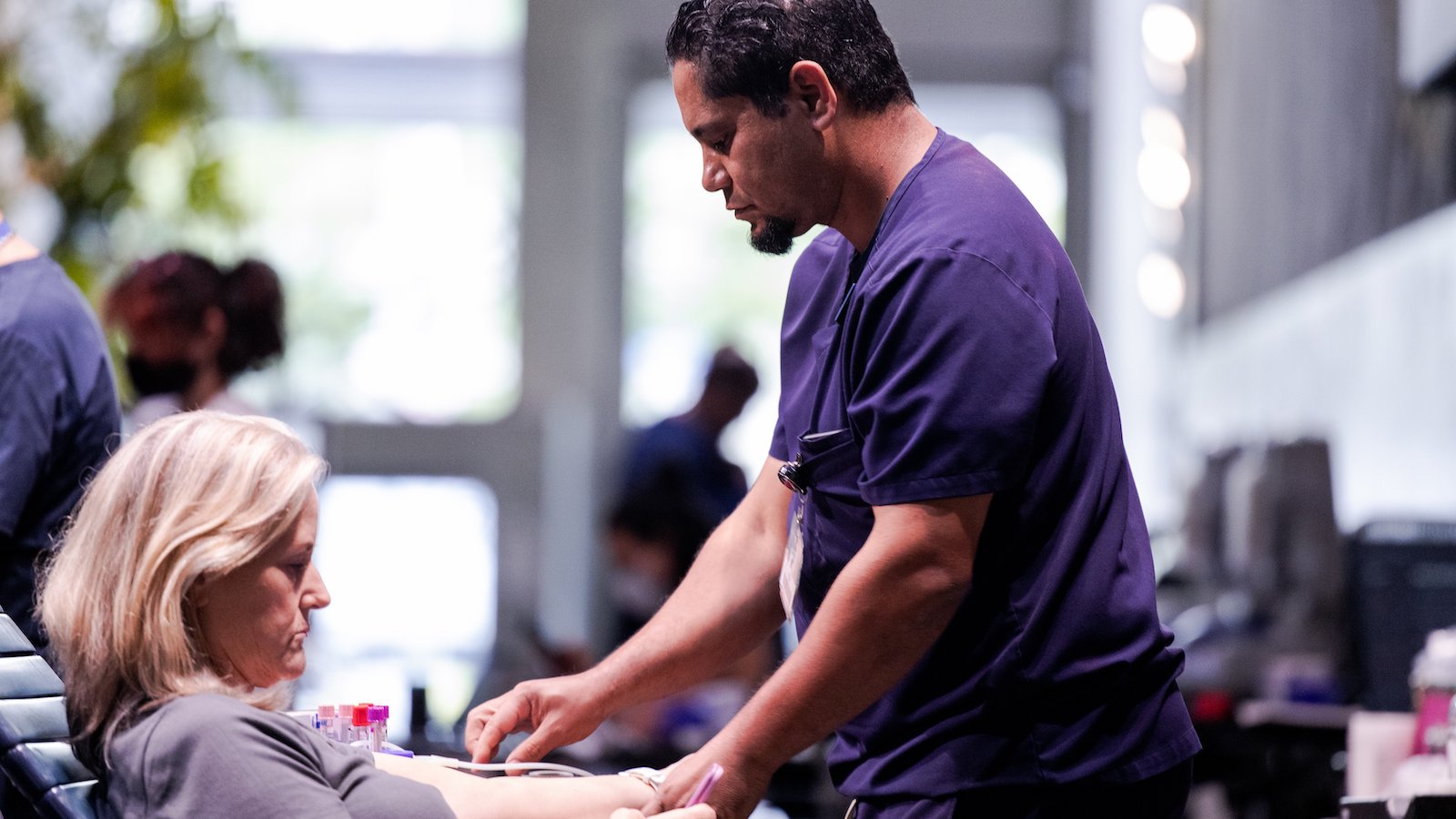 A man with short black hair in dark purple scrubs drawing blood with a woman with short blonde hair