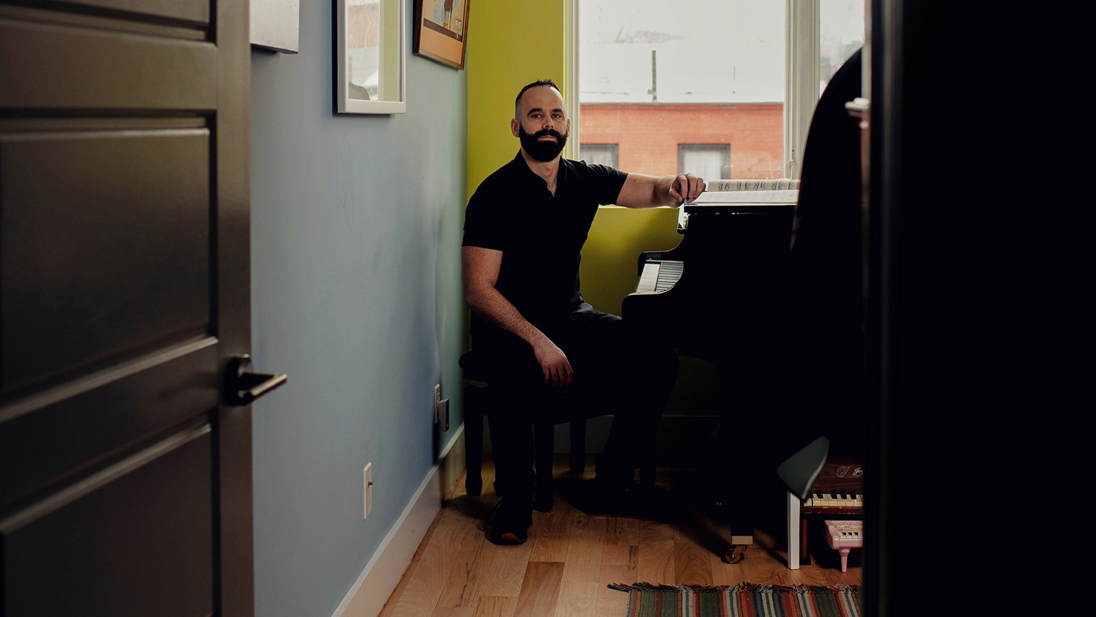 A man with a dark beard and buzzed hair in a black shirt sitting at a piano