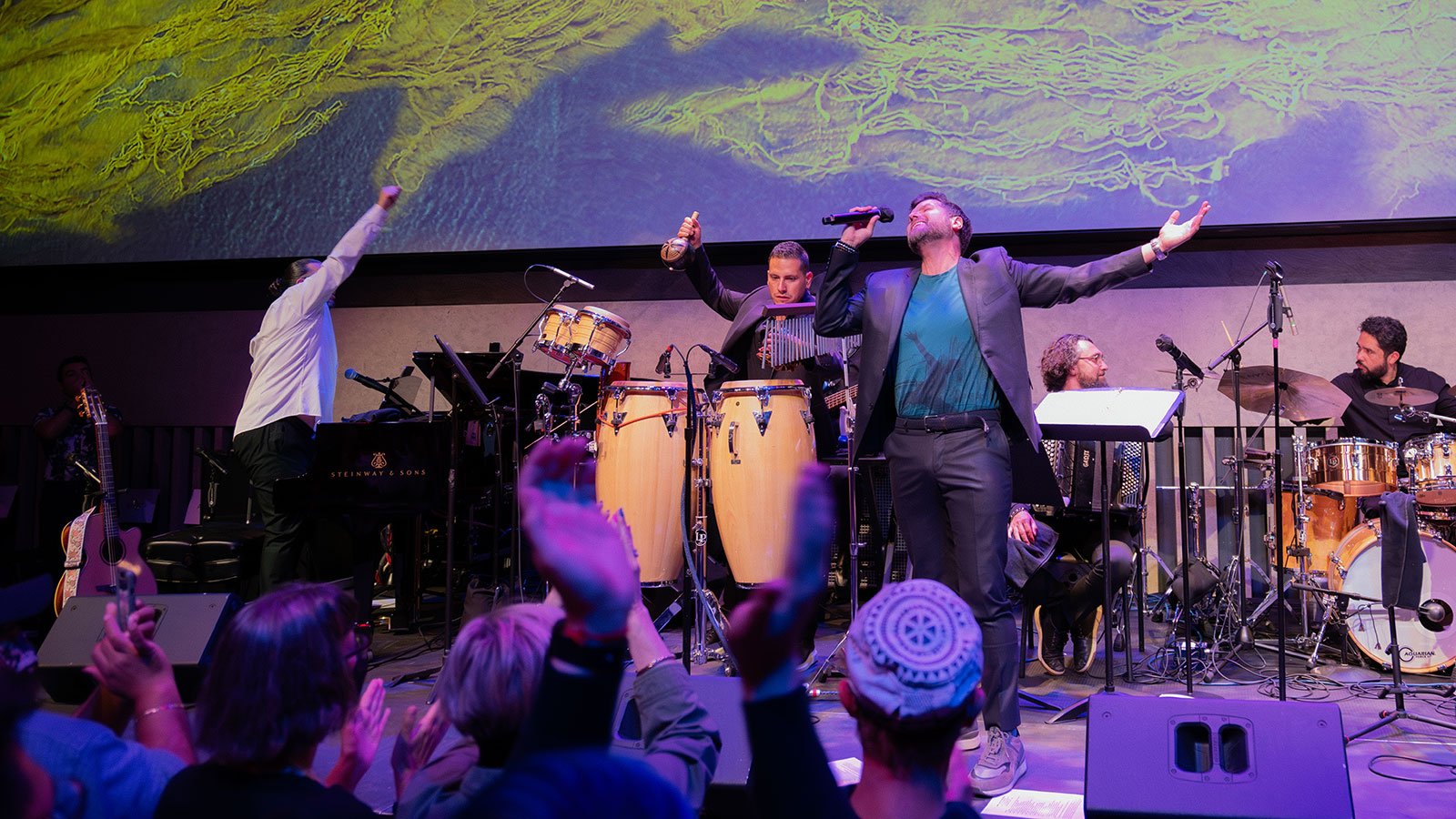 A man in a grey suit and blue t-shirt singing into a microphone with people playing the piano and drums behind him