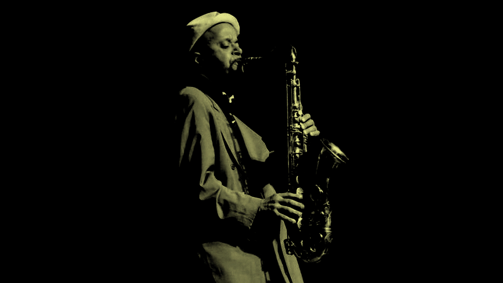 A man in green lighting wearing a long coat playing the saxophone 