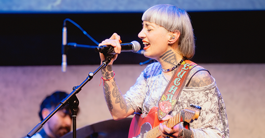 A woman sings into a microphone and holds a red guitar