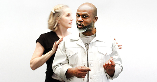 A man stands and looks at the camera and gestures with his hands while a woman behind him holds his shoulders