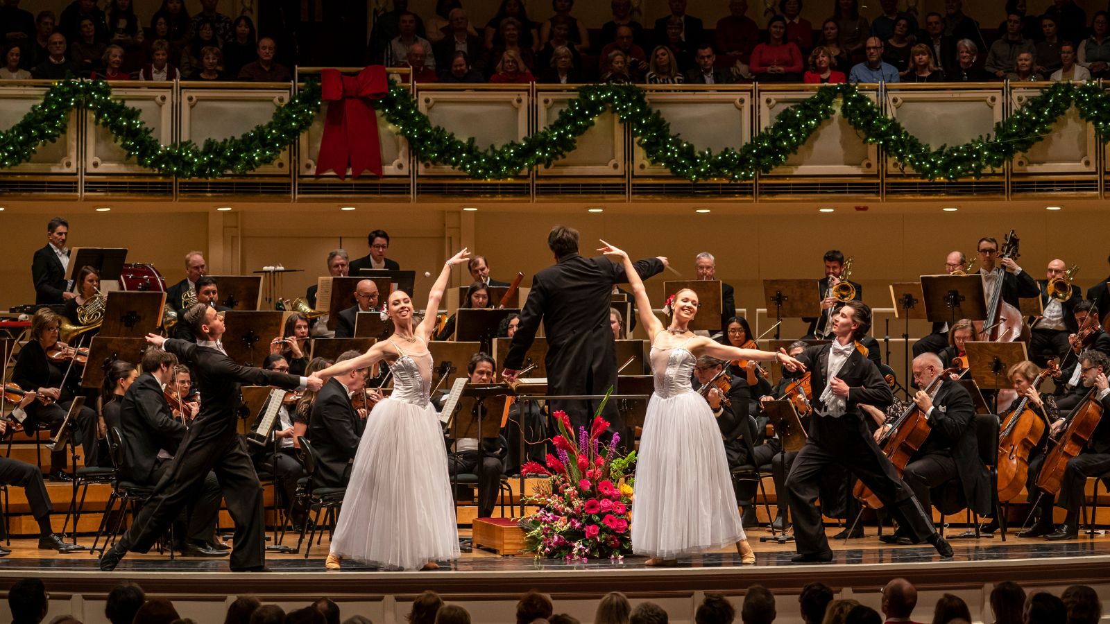 Two dancers in long white dresses in front of a full orchestra