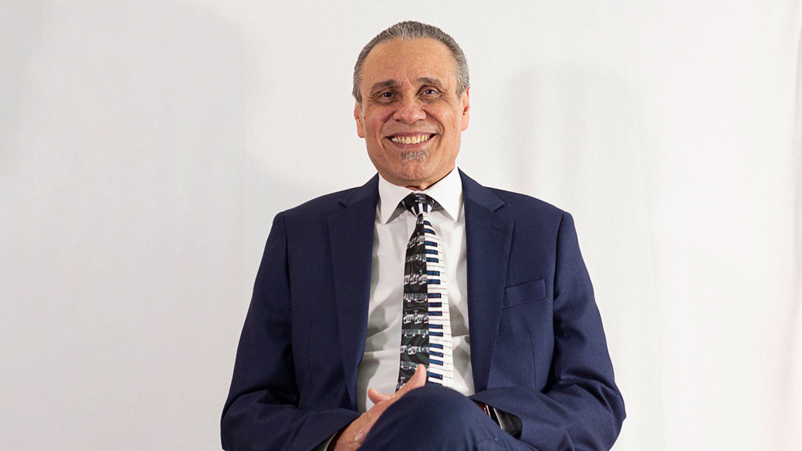 A man with short grey hair wearing a blue suit, white shirt, and a keyboard tie