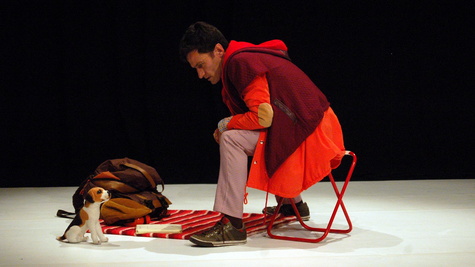 A man in a large red jacket sitting on a red stool looking down at a stuffed dog