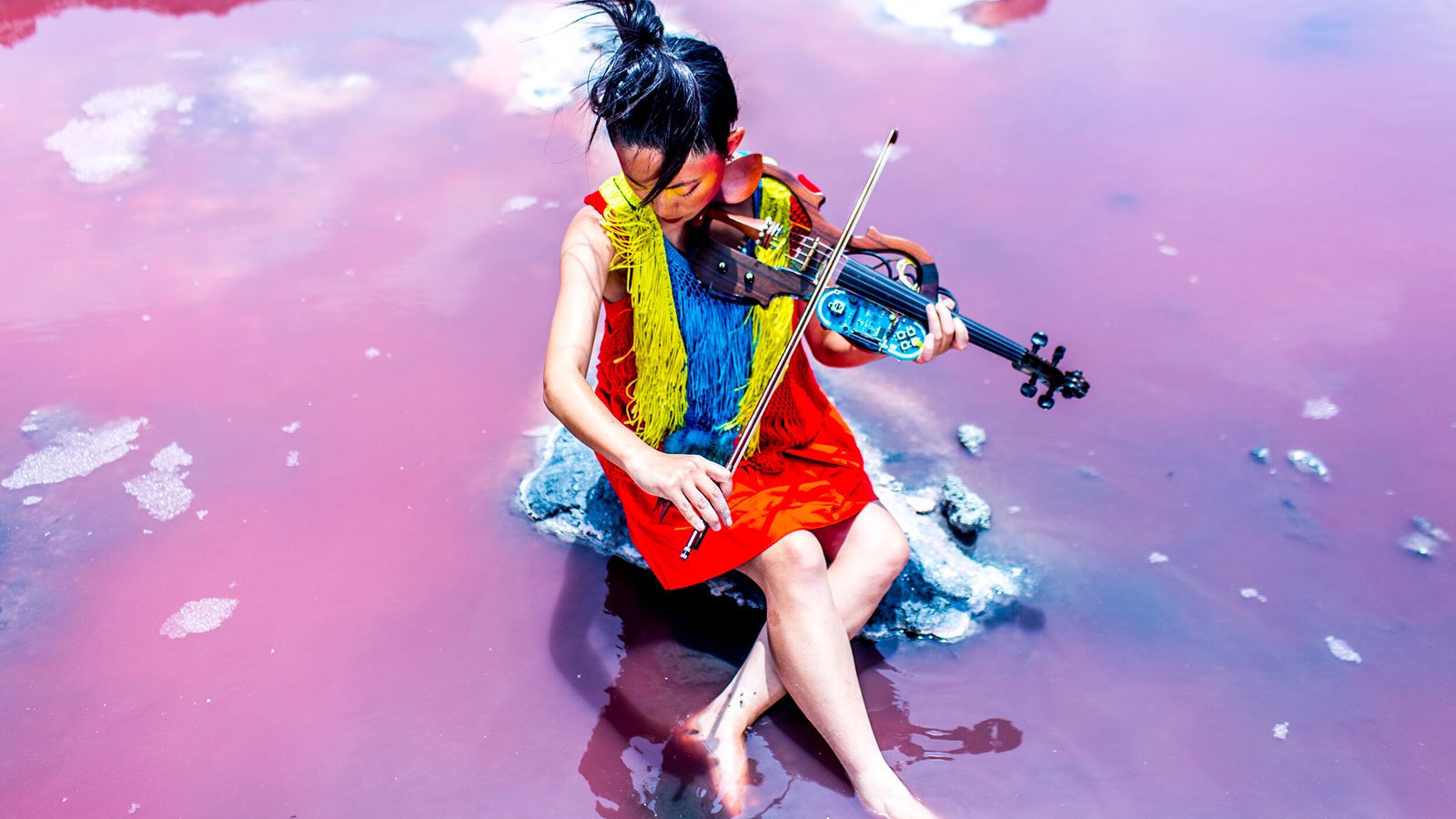 A woman with black hair wearing a red, yellow, and blue dress sitting in water playing a violin