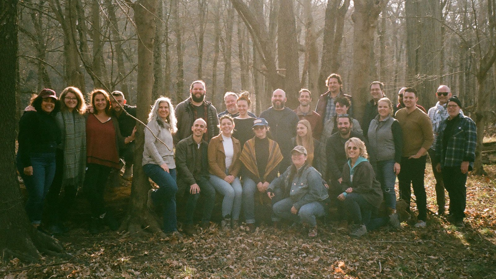 A group of people in fall clothing standing in the woods with leaves on the ground