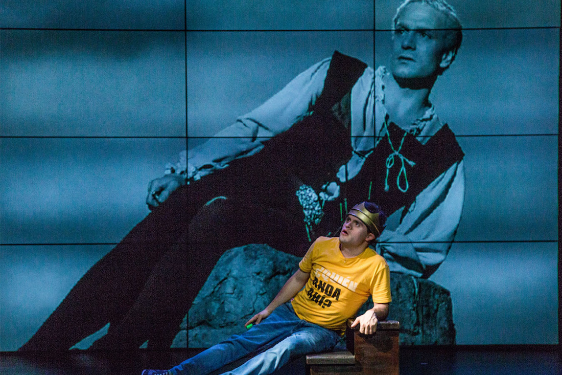 A man in a yellow t-shirt laying on his side in front of a projected version of Hamlet