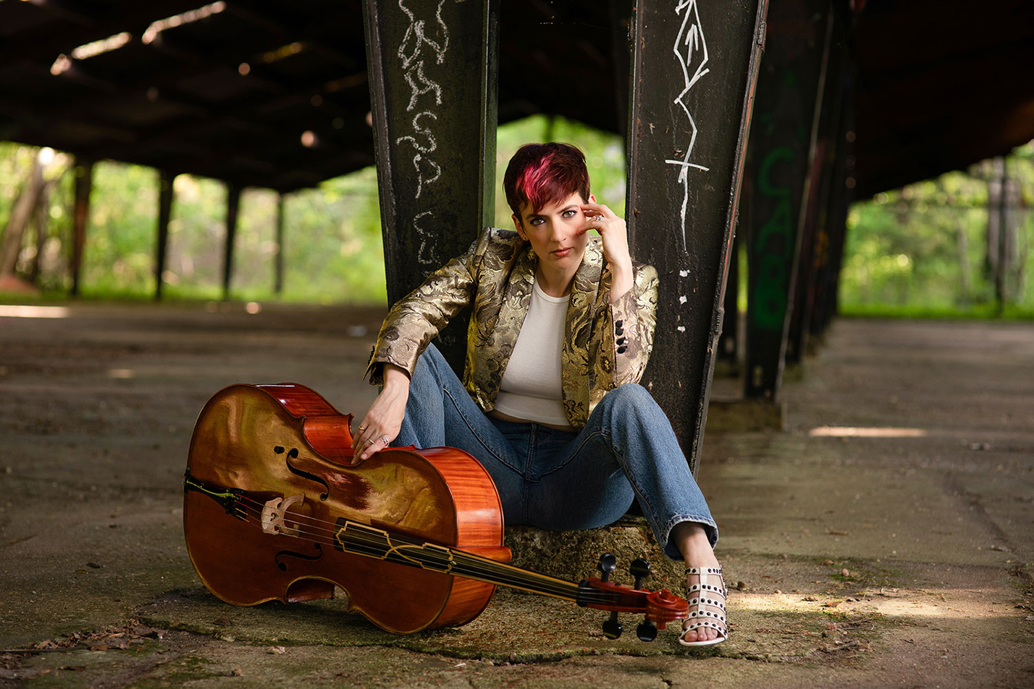 A woman with short red and black hair sitting under an overpass with a cello