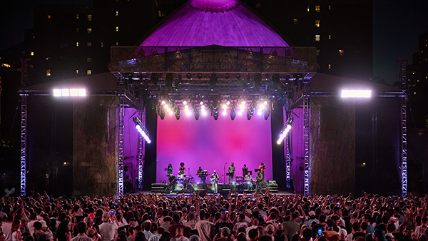 A band plays on the Damrosch Park stage in front of a large crowd.