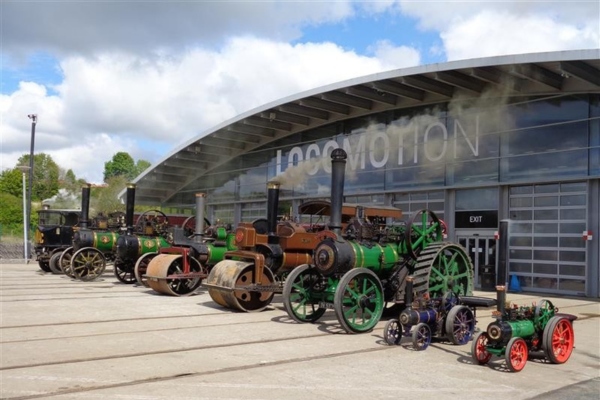 A collection of steam traction engines outside a museum building against a blue sky