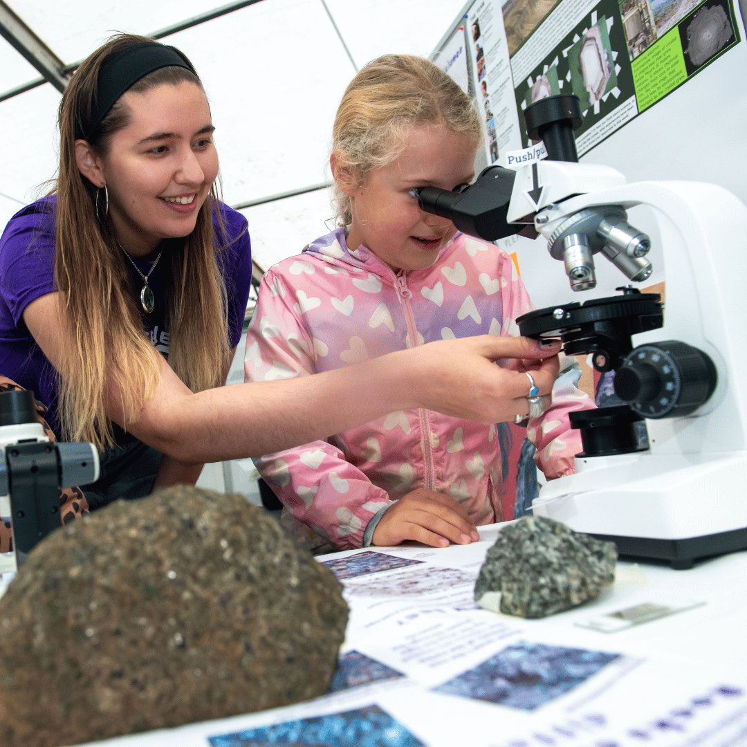 A person with long hair in a purple t-shirt helping a child in a pink coat look through a microscope