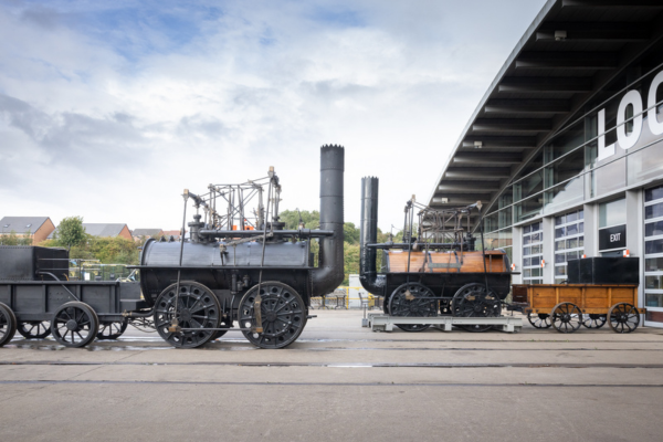 A young girl wearing a green jumper and her grandfather in a navy jumper standing next to 'Locomotion No.1' the original steam engine made from wood and metal