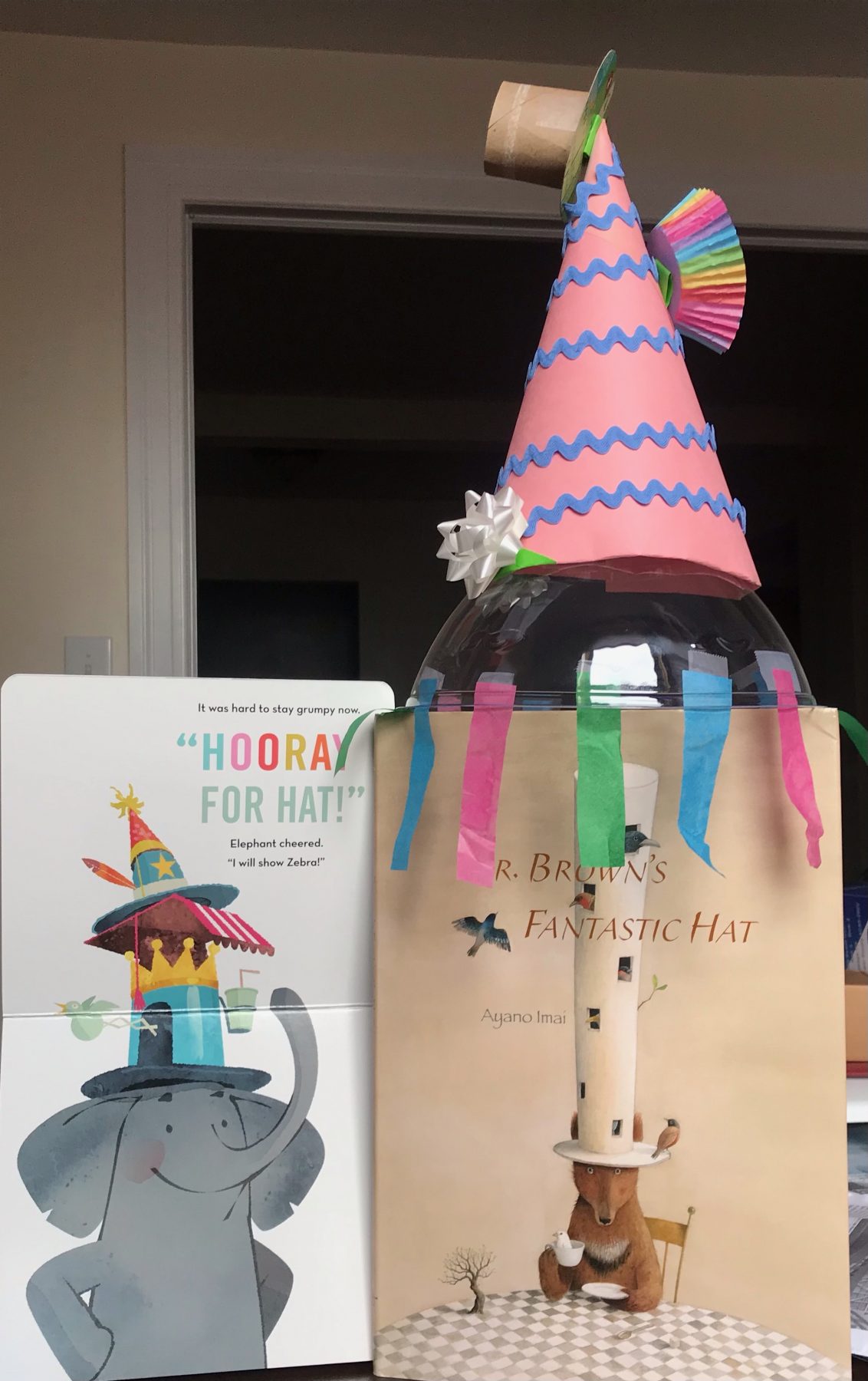 A multicolored hat made of construction paper, streamers, and a rainbow muffin wrapper sits on top of two books.