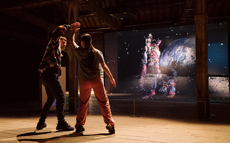 Two dancers interact in front of a screen which displays their 