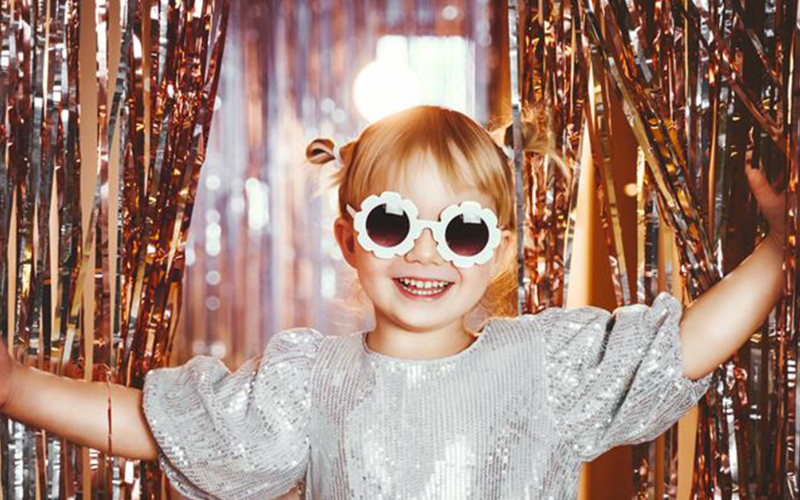 A little girl in flower sunglasses walking through a sequin curtain