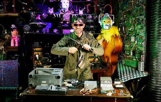 A man in a jumpsuit wearing sunglasses with a table full of old computers and e-waste.