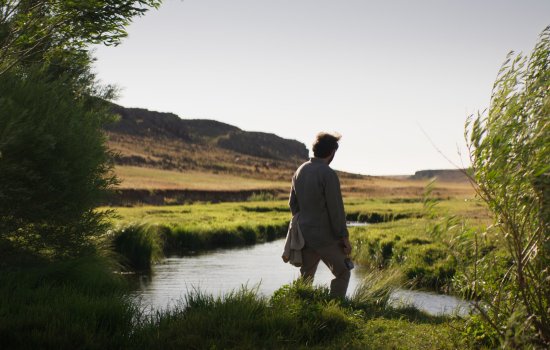 A person in silhouette stands in a river, surrounded by hills and lush greenery.