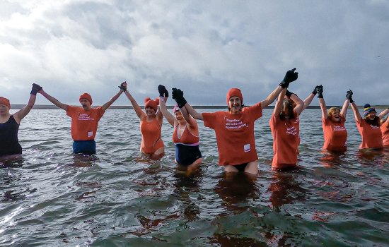 A group of eight people, wearing orange and blue tshirts and swimwear, stand in open water with their arms raised.