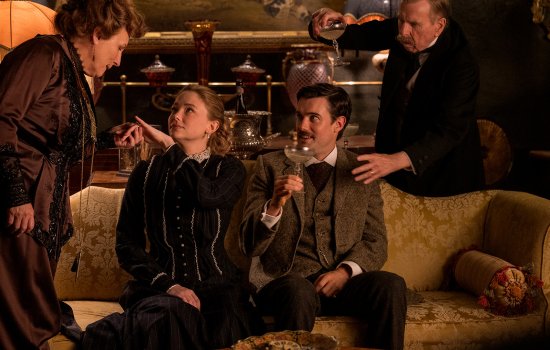 A man and a woman sit on a soft, the man is holding a wine glass. They are looking up at an older woman, behind them a man touches the seated man's shoulder.